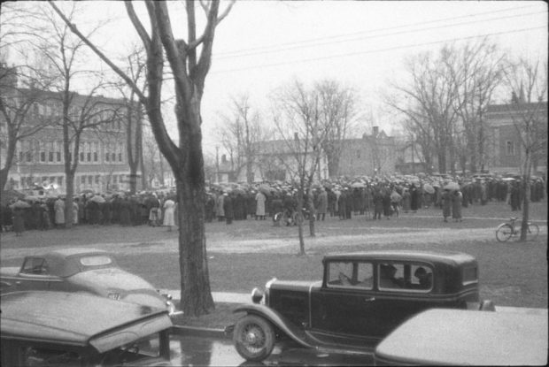 Black and white image of several hundred people with umbrellas gathered in a park, with old cars parked in the foreground.