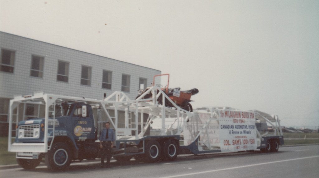 Colour image of an antique car sitting on top of a car transporter in front of an industrial building. A man in a security guard's uniform is standing in front of the car transporter.