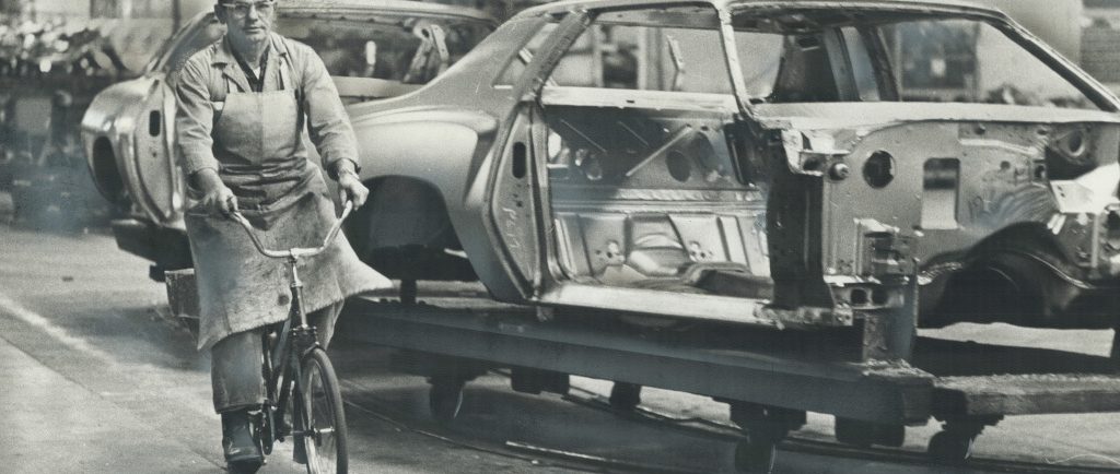 Black and white image of a man in an apron and a backwards baseball cap riding a bicycle alongside an unfinished car on an assembly line.