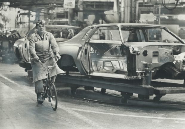 Black and white image of a man in an apron and a backwards baseball cap riding a bicycle alongside an unfinished car on an assembly line.