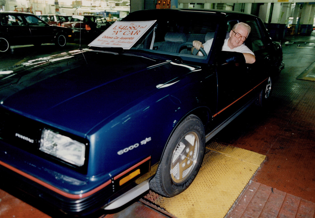 Colour image of a man driving a car through a factory assembly line. A sign on the windscreen reads "The 1,541,882nd "A" Car Oshawa Car Assembly"
