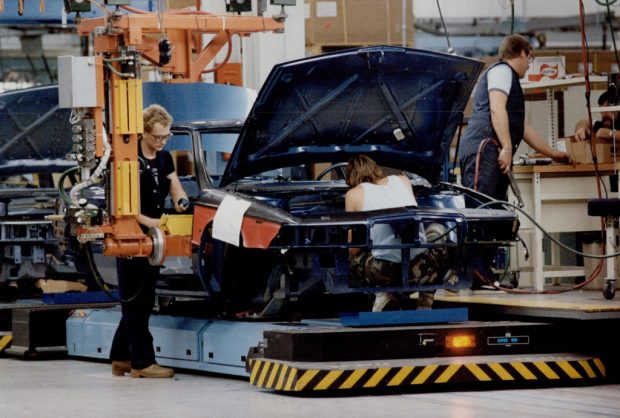 Colour image of three men using heavy machine tools to assemble a partially-completed car on an assembly line.