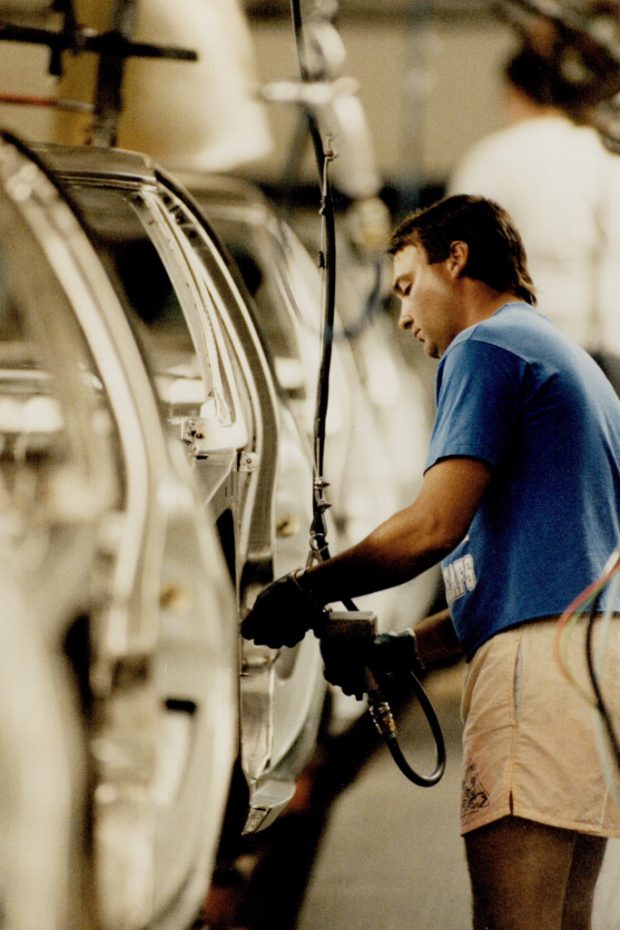 Colour image of a man in a t-shirt and shorts bolting a component into a partially-completed car body.