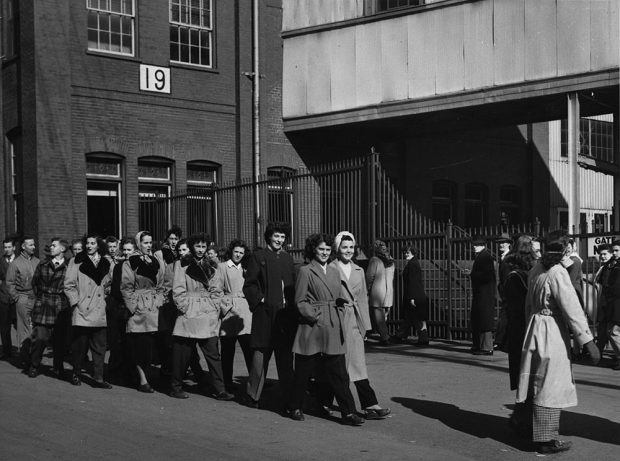 Black and white image of several dozen women and men marching in front of a factory building.