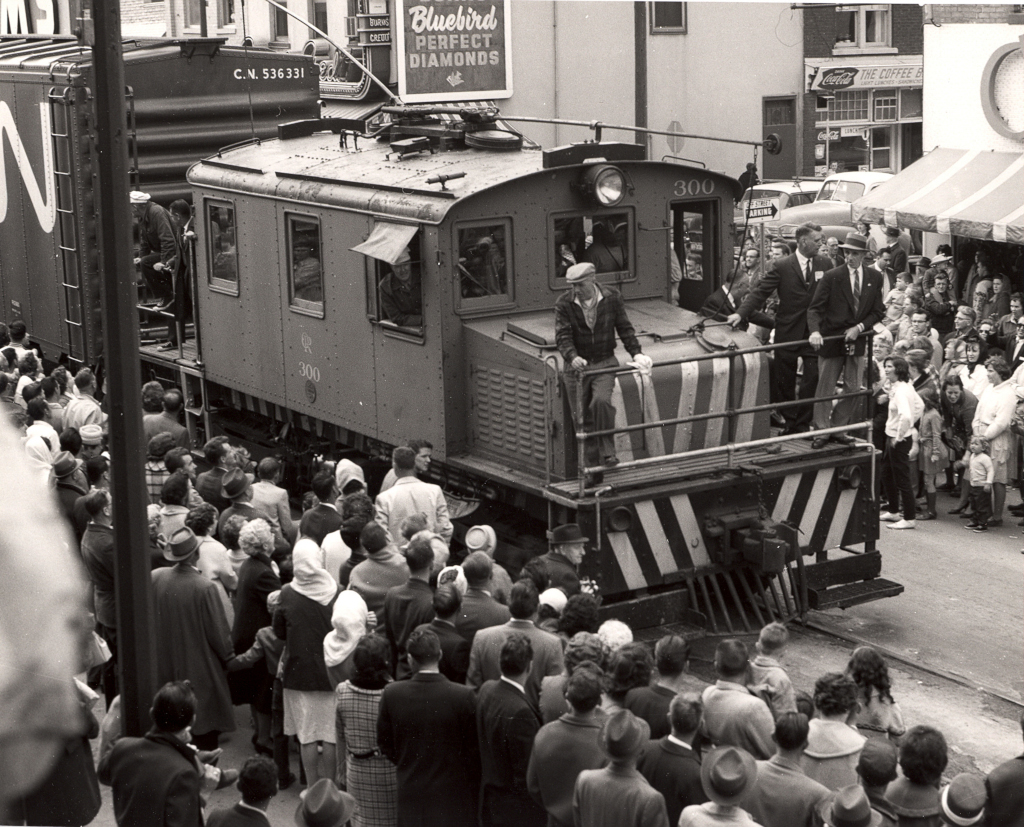 A freight locomotive moves down a crowded street with hundreds of spectators standing on either side.