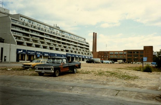 Colour image of a partially-constructed apartment building next to a factory. Several 1970s-era cars and trucks are parked in front of the apartment.