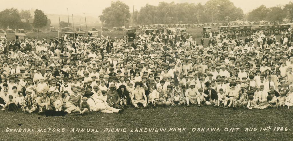 Image of hundreds of people sitting on a lawn in a park. Several dozen automobiles are visible parked in the background. Hand-written image caption reads GENERAL MOTORS ANNUAL PICNIC LAKEVIEW PARK OSHAWA ONT AUG 14TH 1926