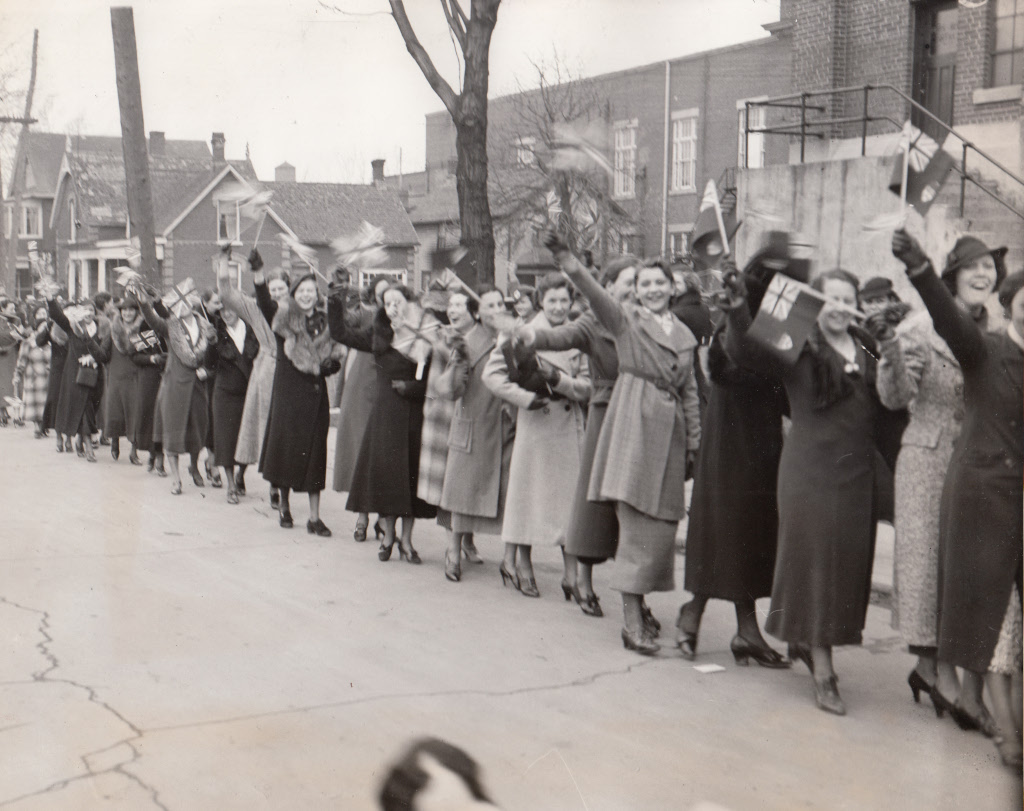 A line of women march in front of an industrial building, waving Canadian and British flags.