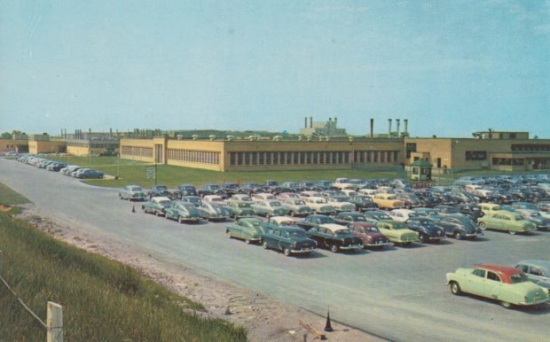 Image of several hundred vintage automobiles parked in front of a factory building.