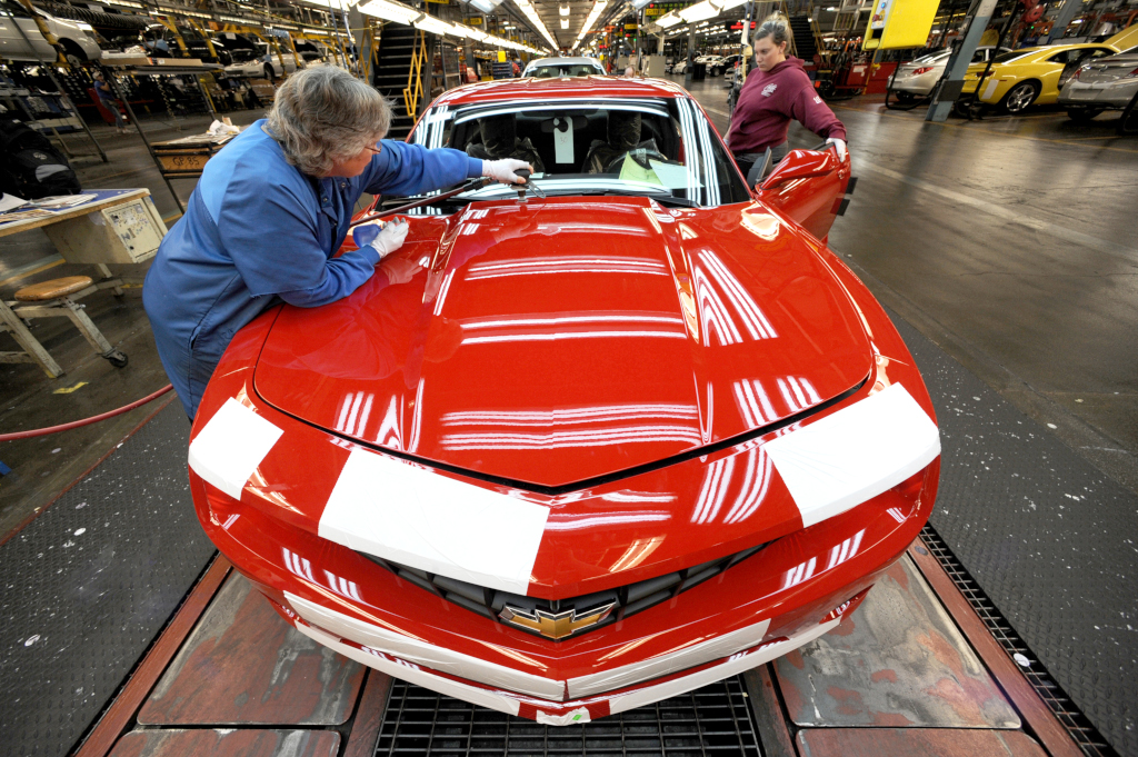 Two women attach parts to a sports car in a factory environment.