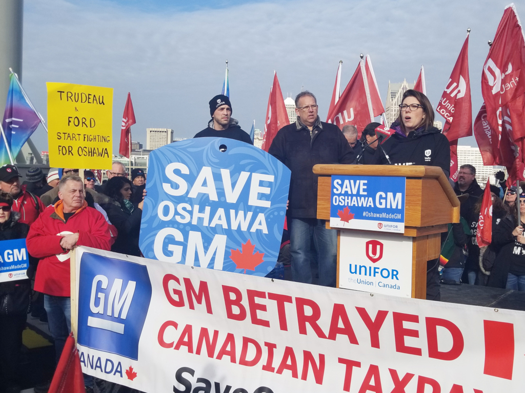 A woman delivers a speech from a podium marked "SAVE OSHAWA GM", while several dozen protesters with flags look on. A hand-written sign in the background reads "TUREAD & FORD START FIGHING FOR OSHAWA"
