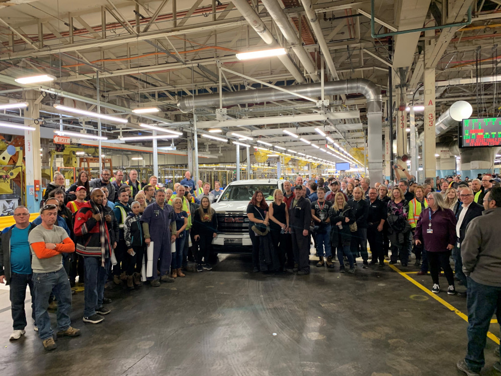 Several hundred men and women, many in work clothes, pose in front of a solitary pickup truck in a factory space.