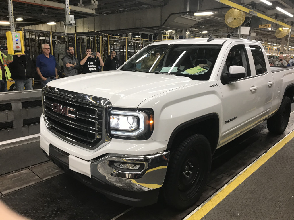 A pickup truck drives through an industrial space. Several men and women stand in the background, taking pictures on cell phones.