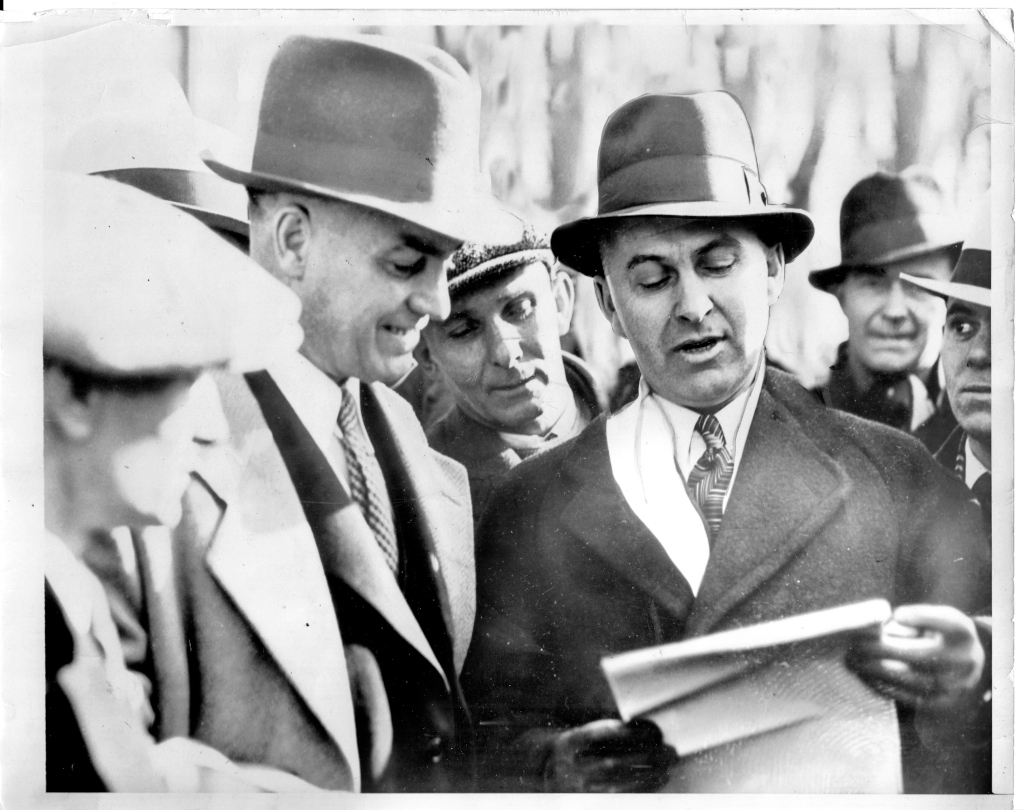 Several men in 1930s clothing discuss the contents of a sheet of paper.