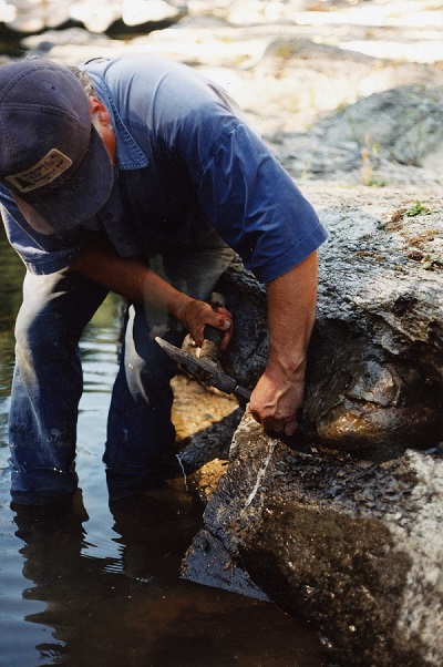 Graham Beard standing over a boulder in the middle of the river, hammer and chisel in hand.