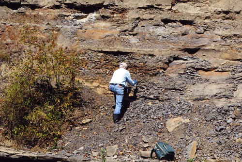 Graham Beard standing against a riverbank wall that displays visible layers of sedimentary rock.