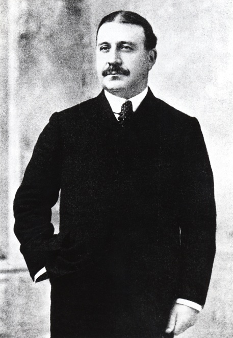 Standing portrait of Francis Clergue wearing a black suit and tie, his hand is in his pocket, his hair is parted down the middle, he has a big bushy moustache.