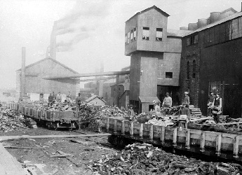 Supplies arriving by steam engine, debris in foreground, workers standing behind train cars.