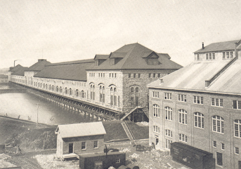 Long sand stone building and two smaller sandstone buildings in the foreground. River to the left of the long building.