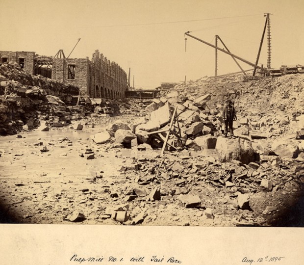 Gravel and stones are strewn across the work site. Cranes, a worker and a half finished building are seen in the background.