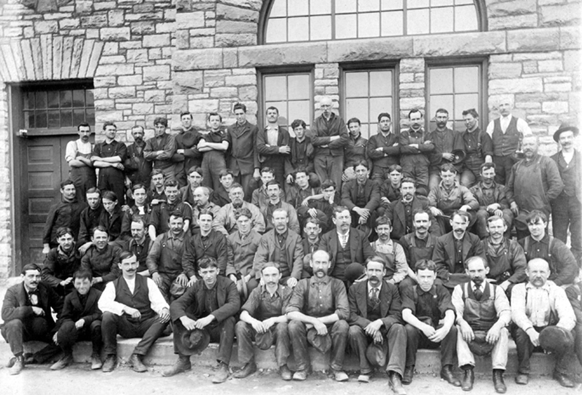 Black and white image of 61 men, sitting or standing in five rows, in front of a stone building.