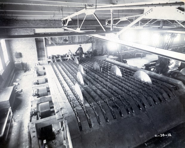 Sawmill machinery, blades facing up, worker overseeing machine.