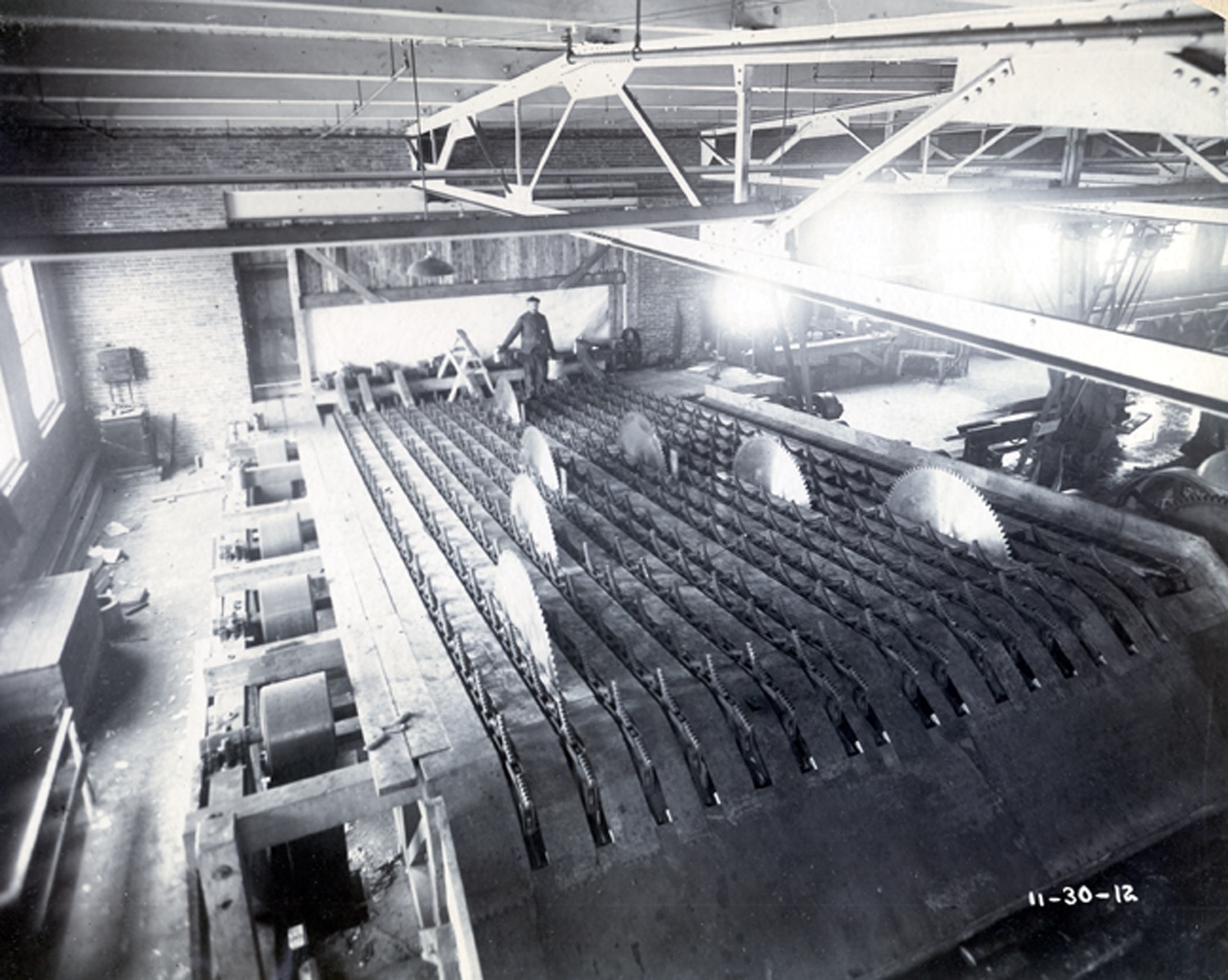 Sawmill machinery, blades facing up, worker overseeing machine.