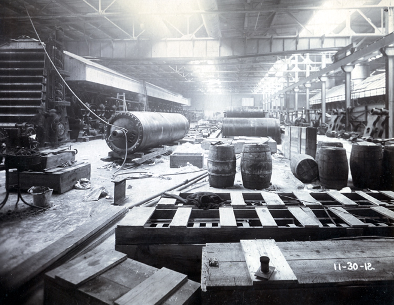 Entrepôt du moulin à papier. Barils et caisses en bois, rouleaux à papier éparpillés sur le plancher.
