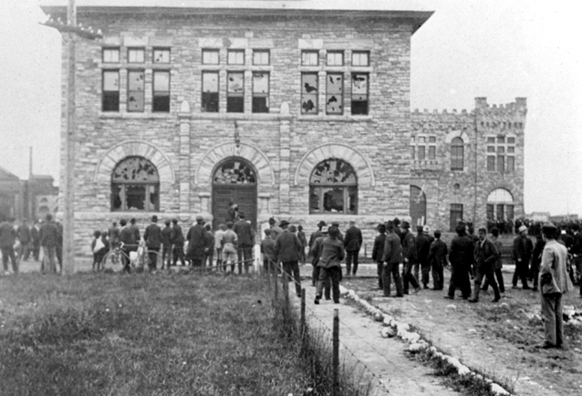 Rioting workers standing in front of sandstone building. The windows of the building have all been broken, presumably by angry rioters.