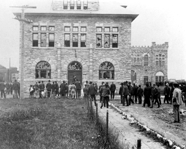 Rioting workers standing in front of sandstone building. The windows of the building have all been broken, presumably by angry rioters.