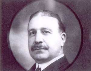 Clergue portrait in circular frame. Dark hair parted in the centre, large moustache. Black suit with white collar.