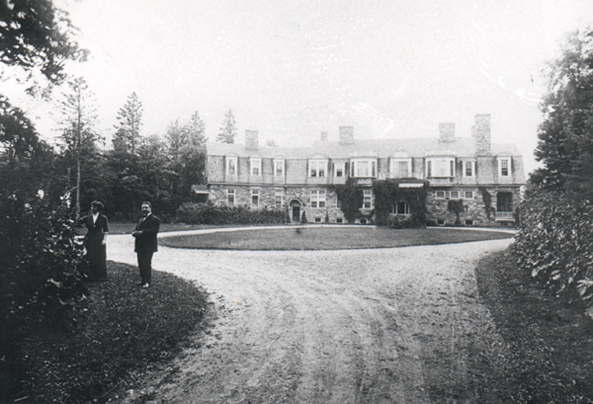 Circular drive way and lawn in front of stone mansion. Two people standing on lawn.