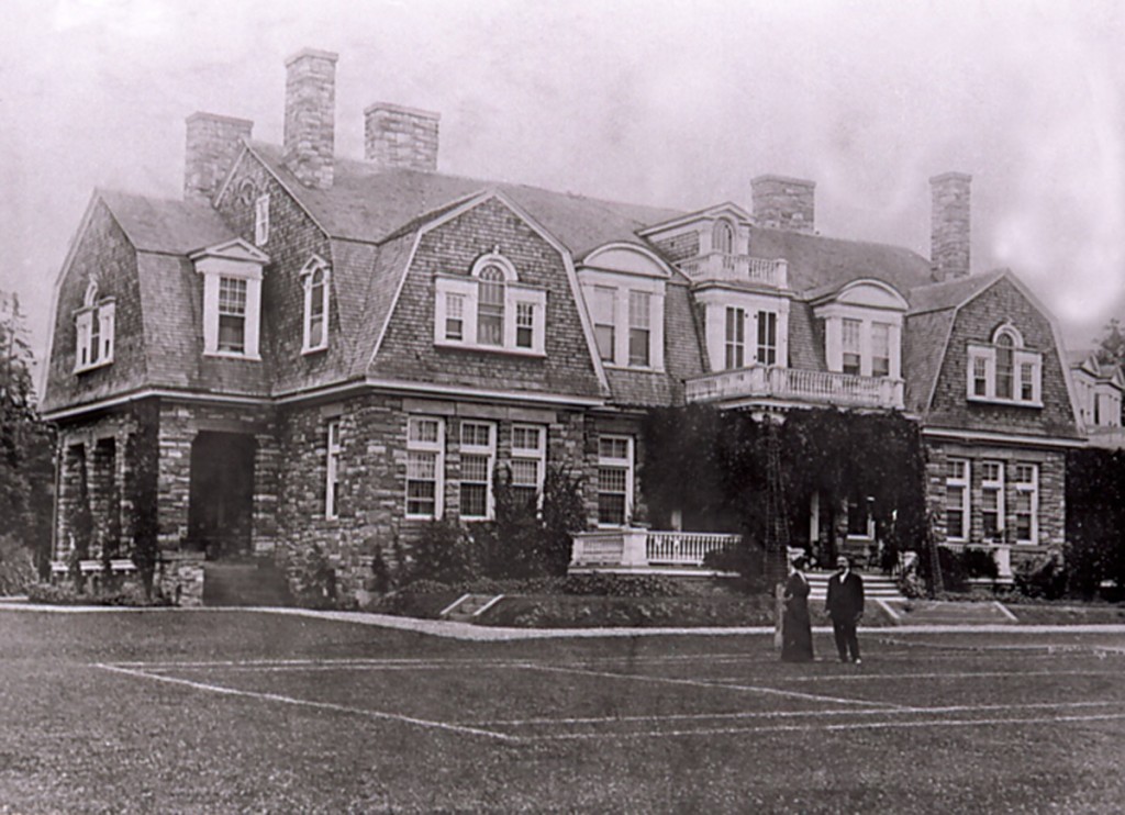 Two people standing on large lawn in front of stone mansion on a hill behind.