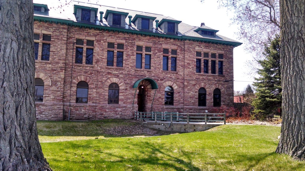Exterior image of the administration building in summer. Three storey sandstone building, with arched windows on the first floor. Green grass in front and bridge/walkway over original Northwest Company canoe lock.