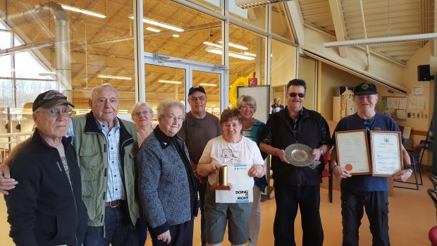 Former employees, predominately seniors, stand together holding paper mill artifacts they collected over the years. Nine people, men and women photographed.