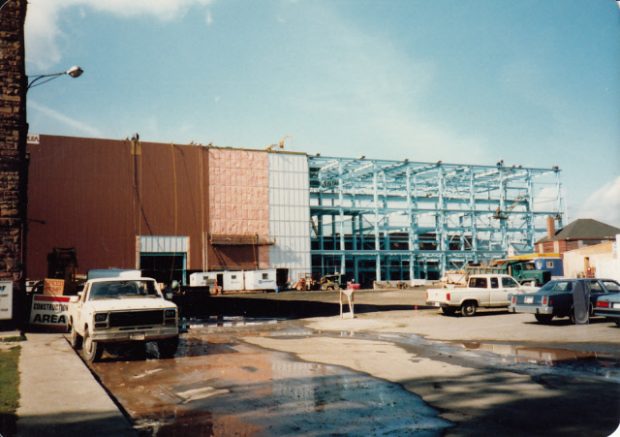 Blue steel beams erected on one side of the new building, the other side to the left is being covered with siding. Trucks are present, a sign leaning against the machine shop reads construction area.
