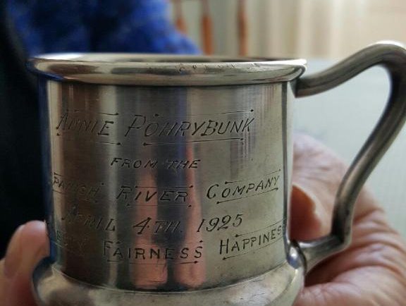 Deux mains de vieille femme qui tiennent une petite tasse en argent portant l'inscription : «To Annie Pohrybunk, April 4, 1925 from the Spanish River Company».