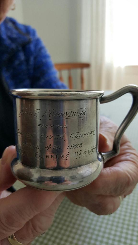 Two elderly hands holding a small silver mug with an engraving to Annie Pohrybunk, April 4, 1925 from the Spanish River Company.