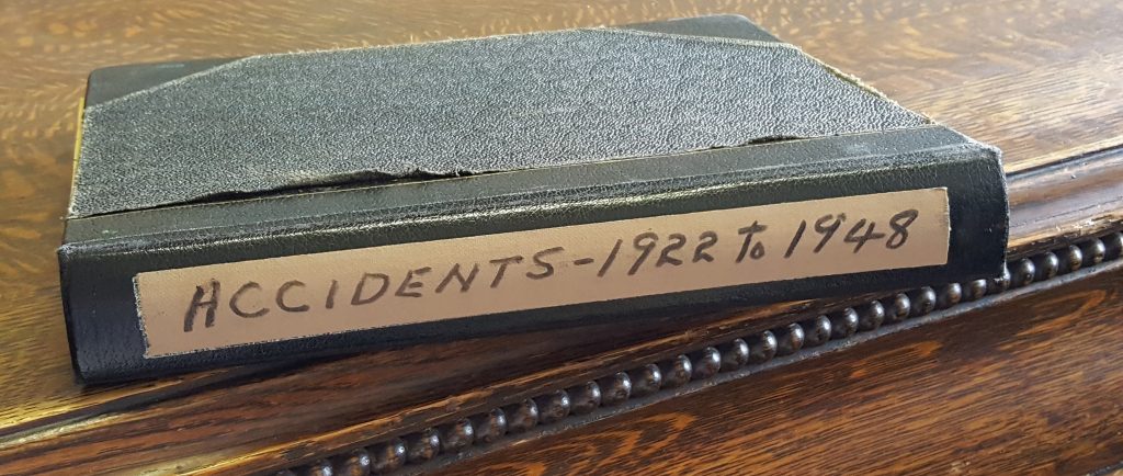 Old black notebook sitting on a wooden table. Tan coloured sticker on the side of the book reads, Accidents - 1922-1948