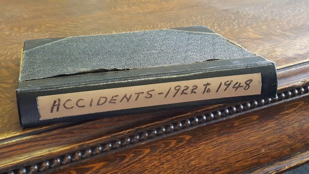 Old black notebook sitting on a wooden table. Tan coloured sticker on the side of the book reads, Accidents - 1922-1948
