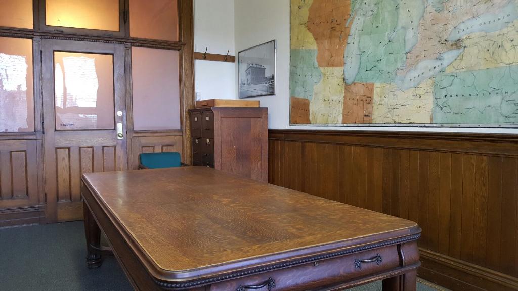 Large wooden desk sitting in the middle of a wood panelled room, view is facing in the direction of the door. Large hand painted wall map is pictured on the right.