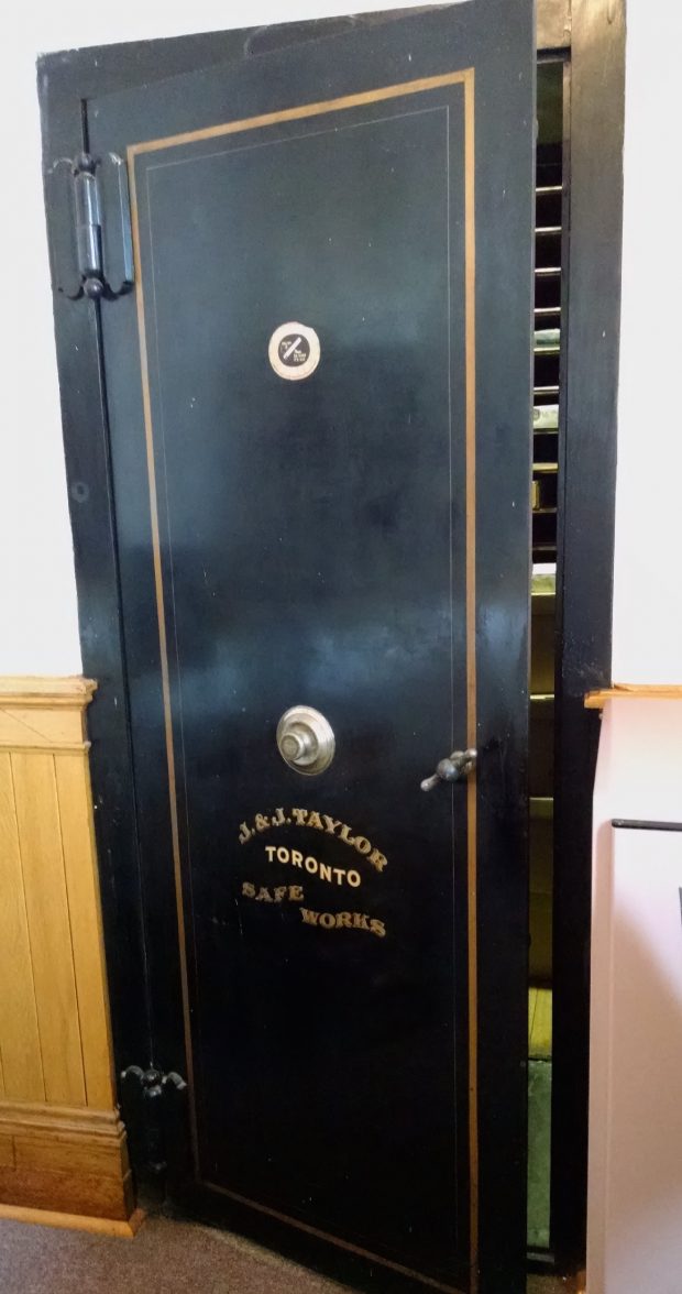 Heavy black metal door of a walk-in safe with a gold painted trim, and J. & J. Taylor, Toronto, Safe Works painted in gold below the combination lock and handle.