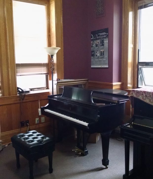Two pianos and a piano bench side by side in a small room that has several big windows, wood panelling, wooden door frames and base boards, is painted burgundy and is carpeted.