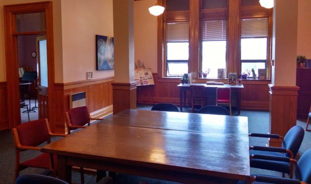 Looking west into the Algoma Conservatory of Music waiting room. View from the door to the room. Big support beams in centre of room. Large window directly to the front. Teaching or office door open on the left of the photograph. Large table and chairs in the centre of the room.