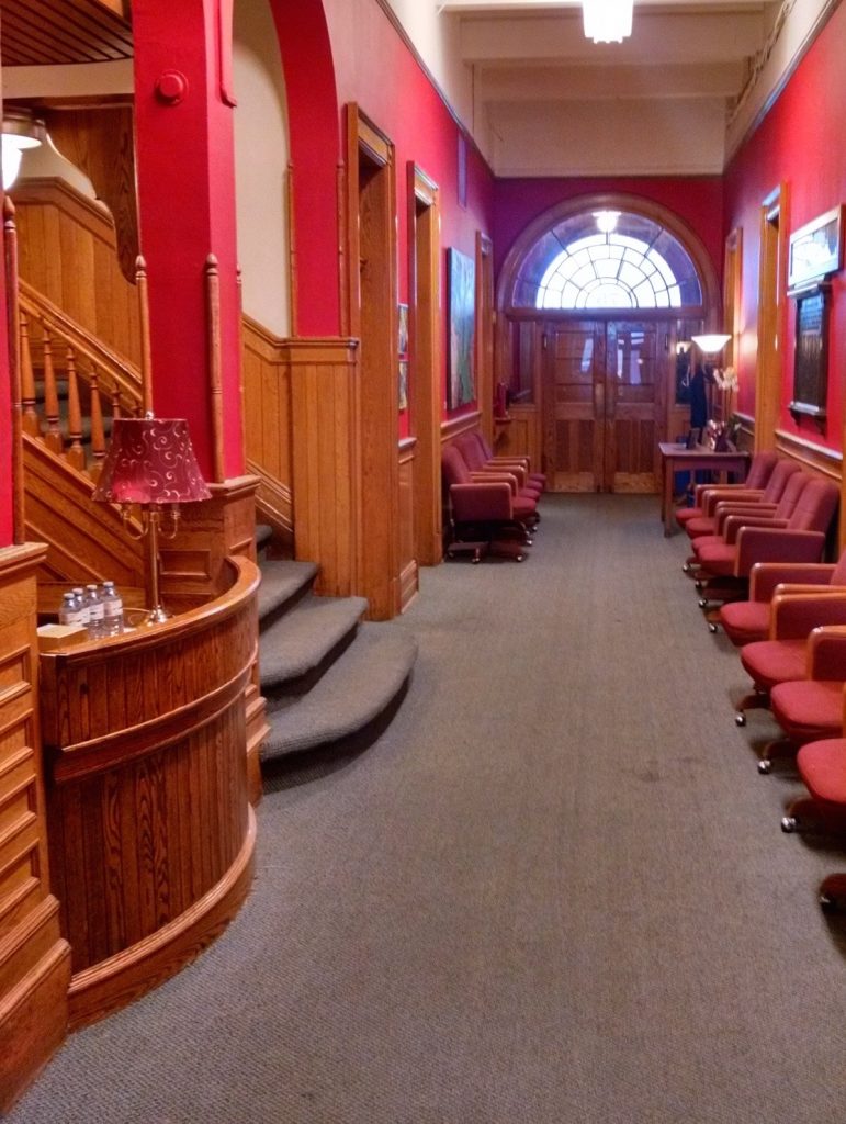 Front entrance and hallway in the Conservatory. Rows of chairs on either side of the hall. Large double doors and windows on one end and stairs and wood detailed reception desk.