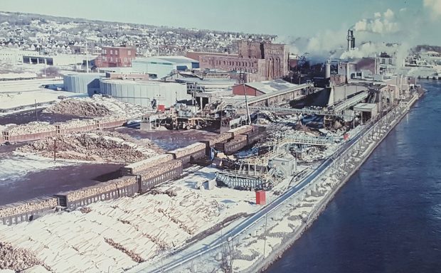 Photo of a framed photograph. Bird's eye view of the paper mill site with buildings, log piles and railway cars