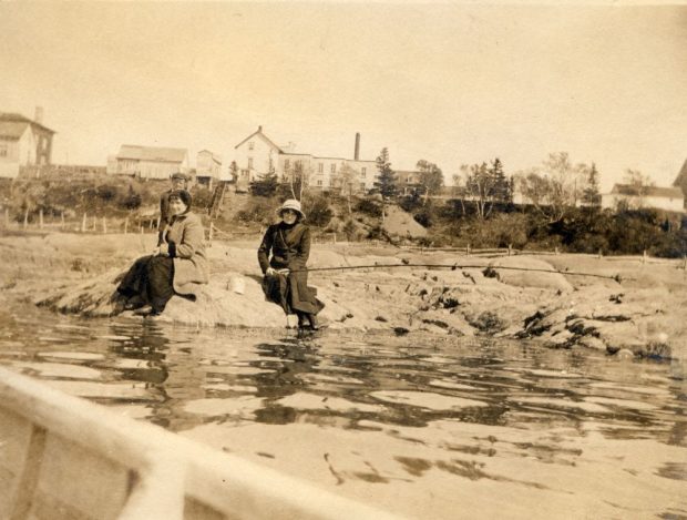 Sepia photograph (around 1910): two women sitting on rocks by the water's edge. Background: shore (grass, trees, buildings). Foreground: part of a boat (photo likely taken from a boat). Coastal/riverside scene.