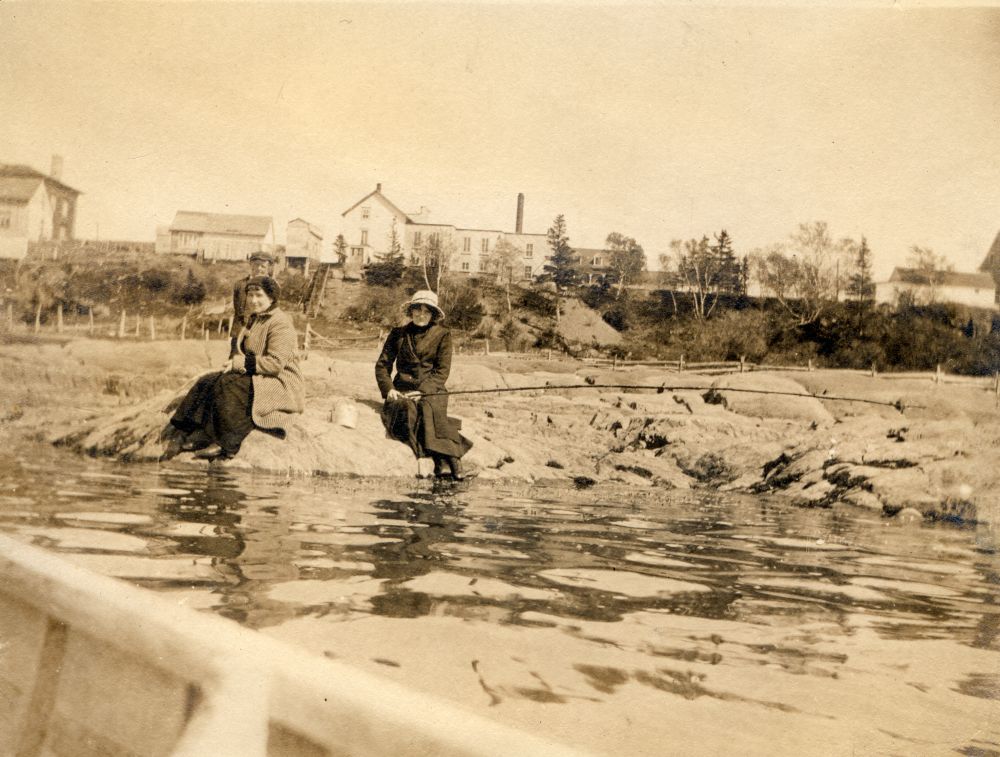 Sepia photograph (around 1910): two women sitting on rocks by the water's edge. Background: shore (grass, trees, buildings). Foreground: part of a boat (photo likely taken from a boat). Coastal/riverside scene.