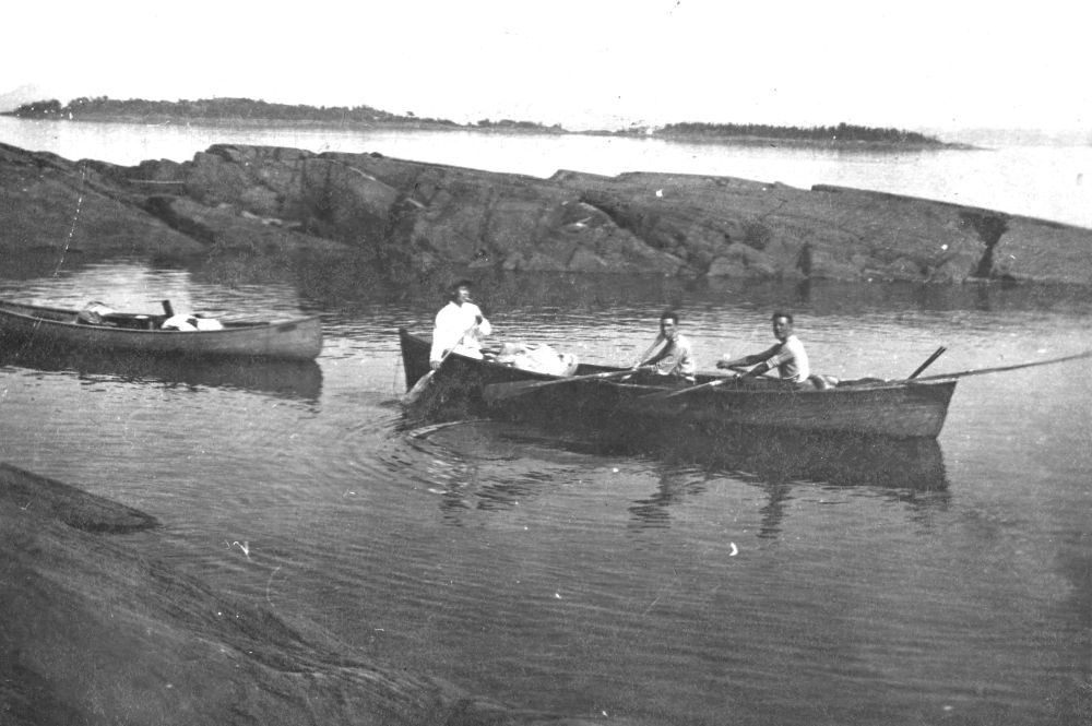 Black and white photograph (1910s): three people in a boat on calm water near a rocky shore. Tight foreground: wooden canoe (three people paddling, light clothing). Left: another canoe. Background: rugged rocky coast, distant islands/land on the horizon. Scene in Kamouraska.