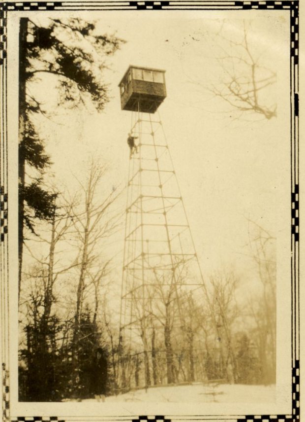 Sepia photograph: tall metal fire tower, winter forest, bare trees. Top of the tower: small square cabin with windows, an observer can be seen climbing the structure. Snow-covered ground, a few conifers. Clear, uniform sky. Decorative border.